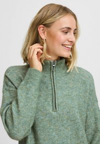 Maglione verde in maglia con colletto alto, dotato di chiusura con mezza zip e polsini a coste. Texture morbida con un sottile motivo mélange.