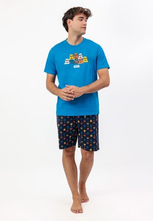 Joven de pie descalzo, vistiendo una camiseta azul de Star Wars y pantalones cortos oscuros con estampados coloridos de cascos de Star Wars.