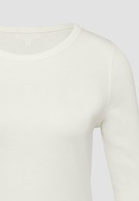 Chemise blanche à manches longues avec col ras du cou, présentant un logo carré en relief subtil sur la poitrine, gros plan montrant la texture du tissu et les détails de la couture.