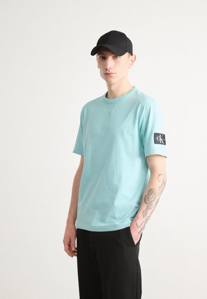 Maglietta di cotone azzurro chiaro con scollatura rotonda e maniche corte, caratterizzata da una toppa nera con testo bianco sul braccio sinistro. Cappellino nero indossato.