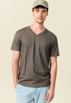 Giovane uomo con maglietta grigia a scollo a V, jeans azzurri e cappellino grigio, in piedi con le mani in tasca davanti a uno sfondo beige.