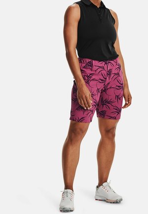 Personne portant un polo noir sans manches, un short rose avec un motif de feuilles noires, et des chaussures de golf blanches à crampons, debout sur un fond blanc.