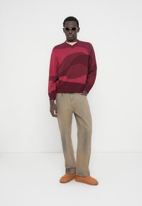 Μπορντό πλεκτό πουλόβερ με αφηρημένο σχέδιο, V-λαιμόκοψη, στενές μανσέτες, μπεζ παντελόνι με φαρδιά μπατζάκια και καφέ παπούτσια τύπου slip-on. Αξεσουάρ περιλαμβάνουν γυαλιά ηλίου.