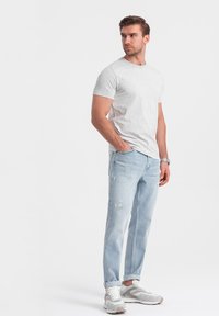 Camiseta de color gris claro con un patrón sutil, combinada con jeans desgastados de color azul claro. El modelo lleva zapatillas blancas y grises. Diseño sencillo.