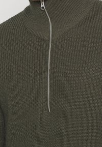 Dunkelgrüner Strickpullover mit hohem Kragen und silberner Reißverschluss, der ein strukturiertes, geripptes Muster und einen lockeren Schnitt aufweist.