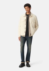 Beige corduroy jacket met twee borstzakken, gelaagd over een zwart shirt met een ruitpatroon. Gedragen met donkerblauwe jeans en zwarte schoenen.