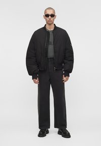 Άνδρας που φοράει μαύρο bomber jacket, σκούρο γκρι γραφικό T-shirt, μαύρο πλατύ παντελόνι τζιν, μαύρα παπούτσια και γυαλιά ηλίου, στέκεται μπροστά σε απλό φόντο.