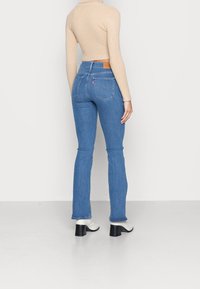 Femme portant un jean bleu clair ajusté, un top court beige à manches longues, et des bottines blanches à talons, debout contre un mur uni.