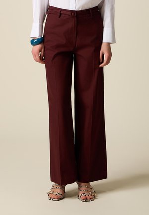 Donna che indossa pantaloni bordeaux a vita alta e gamba larga abbinati a una camicia bianca, bracciale blu e sandali con tacco dal motivo a serpente.