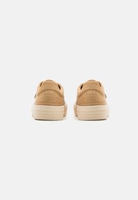 Zapatillas de lona color beige con una parte superior texturizada, diseño sin cordones y suela de yute beige. Presentan costuras sutiles y una forma redondeada.