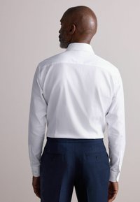 Weißes Langarmhemd mit geradem Kragen, das eine glatte Textur und Knopfmanschetten aufweist, getragen mit navyblauen Hosen.