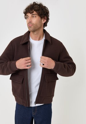 Mand med krøllet hår, der lynlås en mørkebrun jakke over en hvid skjorte, iført blå jeans, står mod en ensfarvet lys baggrund.