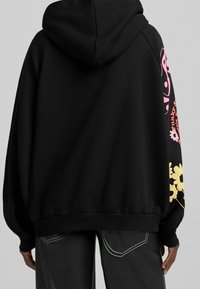 Musta huppari, jossa on värikkäitä kukkakuvioita vaaleanpunaisina ja keltaisina hihoissa, valmistettu pehmeästä ja teksturoidusta materiaalista.