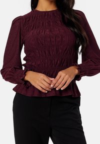 Mujer vistiendo una blusa de manga larga, color burdeos, con fruncidos en los puños y el dobladillo, combinada con pantalones negros, contra un fondo blanco.