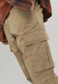 Béžové cargo kalhoty s několika náprsními kapsami, vyrobené z odolné látky, s viditelným šitím. Částečně zakryté hnědou čtvercovou košilí.