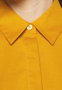 Musztardowa żółta koszula z kołnierzykiem i zapięciem na guziki, charakteryzująca się gładką fakturą i subtelnymi detalami przeszycia z przodu.