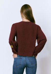 Blouse bordeaux avec des manches longues et bouffantes, et des accents dorés aux poignets. Associée à un jean en denim bleu. Tissu texturé et léger.