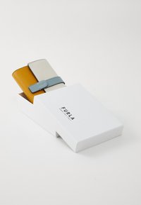 Två läderplånböcker, en senapsgul och en vit med ett blått band, delvis inuti en vit Furla-märkt låda på en vit bakgrund.