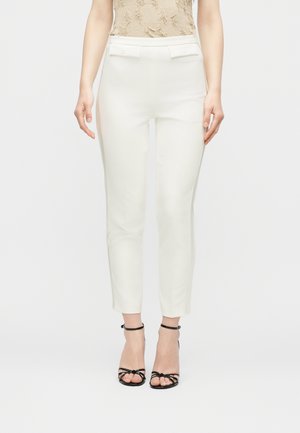 Slim-fit white trousers with decorative belt loops and flap pockets worn with black strappy high-heeled sandals, shown from waist to feet.
