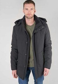 Zwarte geïsoleerde jas met een capuchon, rits aan de voorkant en zakken. Heeft een gladde textuur en een relaxte pasvorm. G gedragen over een groene shirt.