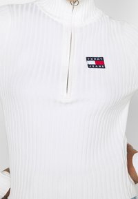 Haut blanc côtelé avec fermeture éclair à quart, arborant une petite écusson logo Tommy Jeans en noir, rouge et blanc sur le côté gauche de la poitrine. Texture lisse.