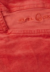 Korallfärgad kordflbased tyg med en mjuk textur, prydd med en broderad logotyp i matchande ton. Sytt i kanterna och subtila vertikala ribbdetaljer.
