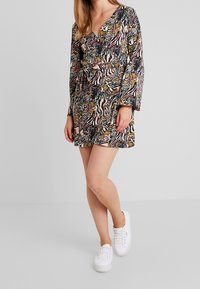 Kwiatowa sukienka typu wrap z długimi rękawami, ozdobiona czarnym, białym i złotym wzorem zwierzęcym. Połączona z białymi sneakerami dla uzyskania casualowego wyglądu.