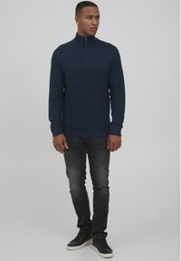 Suéter de punto azul marino con cremallera y un patrón texturizado, combinado con jeans gris oscuro y zapatillas negras. Presenta un cuello alzado y puños de canalé.