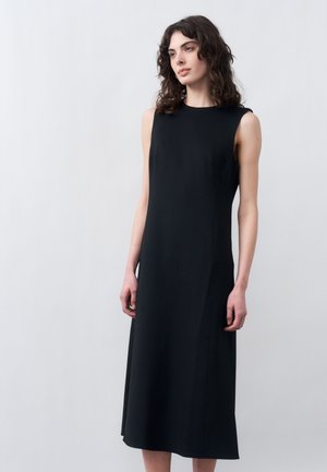 Femeie cu păr ondulat și întunecat, purtând o rochie neagră fără mâneci, până la genunchi, stând în picioare pe un fundal simplu, gri deschis.