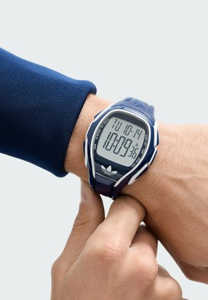 Digitālā pulkstenis ar tumši zilu silikona siksniņu un baltām detaļām. Tam ir taisnstūra forma, liels digitālais displejs un datuma/laika indikators.