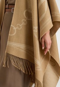 Poncho in lana beige con orlo frangiato, caratterizzato da un sottile motivo a catena. Texture morbida e design ampio per sovrapporsi all'abbigliamento.
