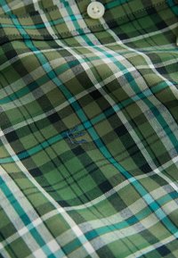 Green plaid shirt made of fabric with a smooth texture. Features turquoise, black, and white lines, and a small embroidered logo.