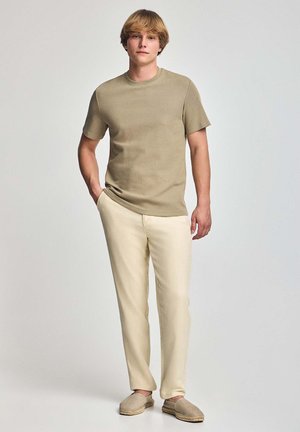 Camiseta beige de manga corta con escote redondeado, combinada con pantalones color crema claro y zapatos sin cordones beige, sobre un fondo gris.
