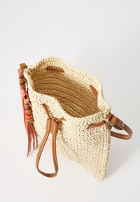 Kudottu olkilaukku ruskeilla nahkadetaileilla, sylinterimäinen muoto ja pehmeä pinta. Varustettu nyöri sulkimella ja värikkäillä tupsuilla.