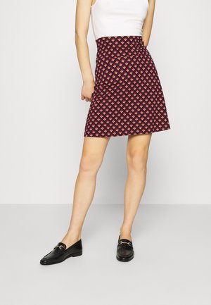 Woman wearing a red and black patterned knee-length skirt, white sleeveless top, and black loafers standing against a plain wall.