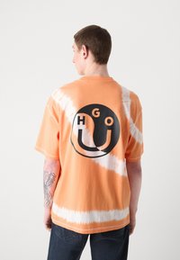 Trumpetakinės rankovėmis oranžinės ir baltos spalvų "tie-dye" marškinėliai su dideliu juodu apvaliu logotipu ir užrašu "HGO U" ant nugaros. Pagaminti iš medvilnės, laisvas kirpimas.