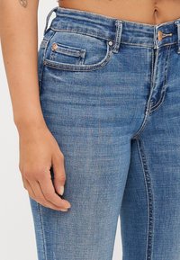 Siniväriiset farkut, joissa on korkea vyötärö, etutasku, kuparinväriset yksityiskohdat ja kevyt haalistuminen kankaan tekstuurissa.