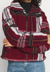 Veste en polaire à carreaux rouges avec un col haut, fermeture éclair devant et rayures contrastantes en bleu, blanc et marron. Tissu texturé et doux.