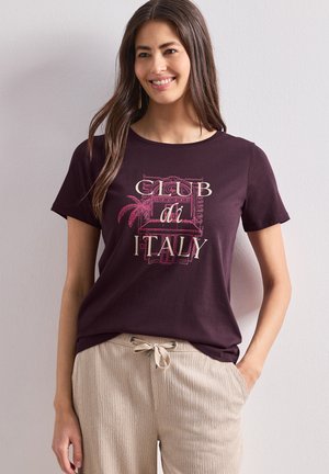 Femme souriante aux longs cheveux foncés porte un t-shirt violet foncé avec l'inscription « Club di Italy » et un pantalon beige à cordon.