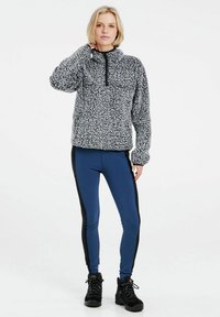 Fleece-Pullover im Schwarz-Weiß-Muster, mit einem Viertel-Reißverschluss auf der Vorderseite; kombiniert mit blauen Leggings mit schwarzen Seitenstreifen und schwarzen Wanderschuhen.
