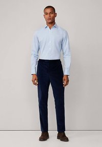 Camicia azzurro chiaro con bottoni e tessuto liscio, abbinata a pantaloni di velluto a coste blu scuro. Il modello è in piedi, indossa stivali marroni.