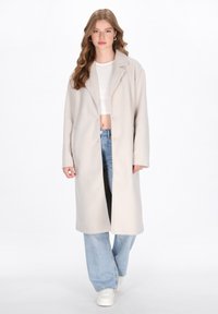 Cappotto lungo beige con colletto a punte, due bottoni e tasche laterali. Indossato sopra un crop top bianco e jeans a gamba larga blu. Texture liscia.