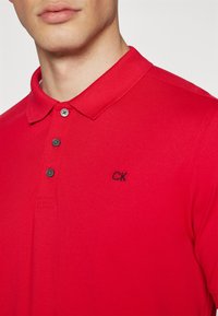 Crvena polo majica s teksturiranim materijalom, klasičnog ovratnika i tri gumba. Crni vezeni "CK" logo na lijevoj strani prsnog koša.