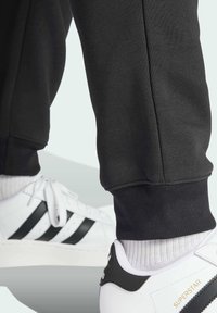 Sneaker Adidas Superstar bianche con strisce nere indossate con calze bianche e pantaloni tuta neri arricciati alla caviglia.