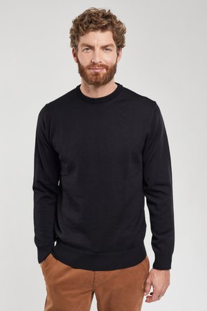 Man met krullend haar en baard, gekleed in een zwarte trui met lange mouwen en een bruine broek, staand met handen in de zakken.