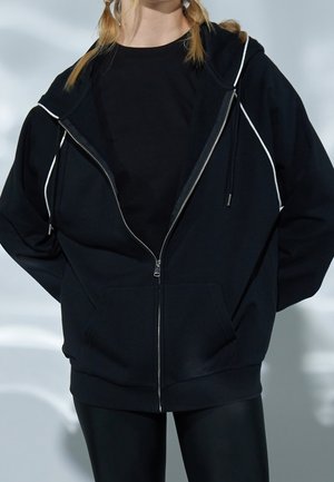 Felpa con zip nera con accenti bianchi, vestibilità oversize, polsini a costine e grande tasca anteriore. Abbinata a leggings neri.