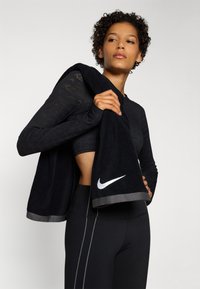 Fekete, hosszú ujjú póló, texturált mintával, fekete, magas derekú leggingsszel párosítva. Egy sötét törölköző, amelyen a Nike logó látható, az egyik vállon lóg.