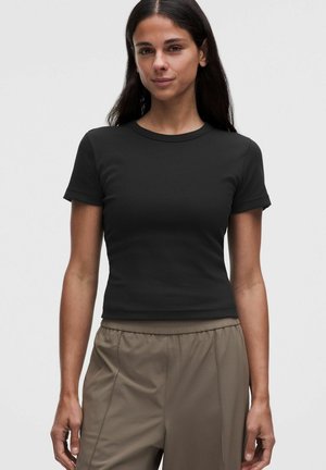Donna con lunghi capelli scuri che indossa una maglietta nera attillata a maniche corte e pantaloni larghi marrone chiaro su uno sfondo grigio uniforme.