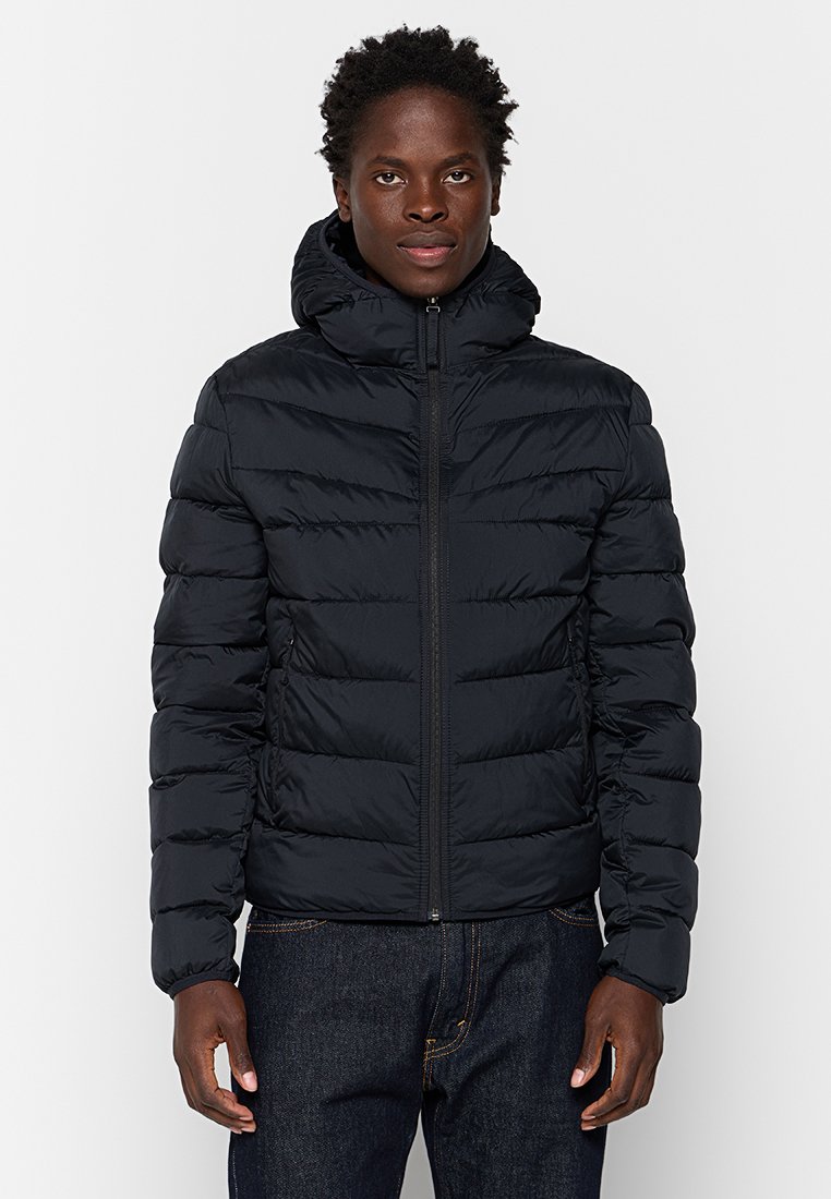 Czarna puchowa kurtka z zapięciem na zamek i kapturem. Posiada pikowany wzór i elastyczne mankiety. Noszona z ciemnoniebieskimi dżinsami.