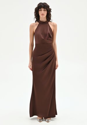 Femme aux cheveux bouclés portant une robe sans manches, à col montant, longue jusqu'au sol, de couleur marron foncé avec des détails drapés et des chaussures métalliques pointues.